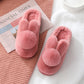 Burgundy plush bunny slippers with ears and pom pom tails lying on a white knit blanket beside a newspaper