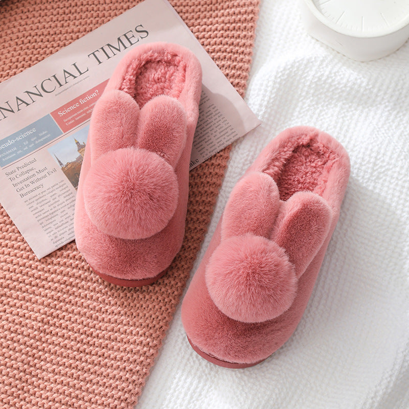 Burgundy plush bunny slippers with ears and pom pom tails lying on a white knit blanket beside a newspaper
