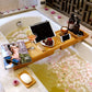 Natural bamboo bathtub caddy tray across a tub with a tablet, a pink drink, fruit, a candle and a magazine