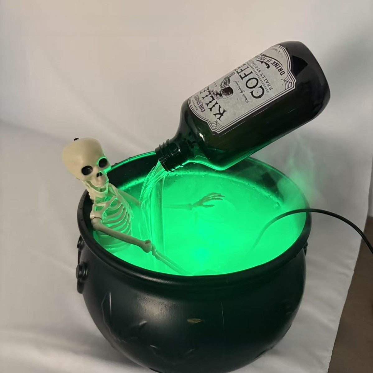 Small black cauldron glowing green with a skeleton sitting in the pot and a bottle pouring a green stream