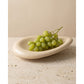 Glossy beige ceramic oval fruit bowl holding green grapes on a travertine surface