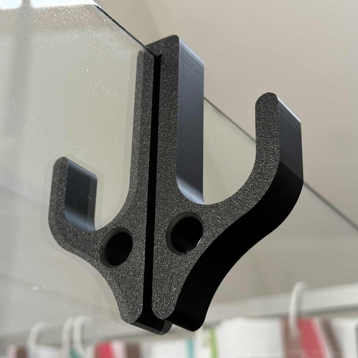 Black resin double hook with two round holes sitting over the top edge of a glass shower door