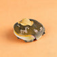 Olive resin frog earbud case wearing a woven straw hat, closed, on a peach background