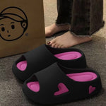 Black EVA heart cutout slide sandals beside a paper bag on a grey floor