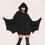 Woman in a black oversized hoodie with arms spread so the bat wing sleeves open, striped tights, pale pink background