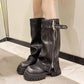 Black chunky platform knee high boots with buckle strap and side zipper worn on legs against a beige wall
