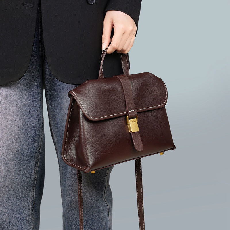Woman holding the coffee brown flap satchel by its top handle, gold-tone buckle on the front, strap hanging down