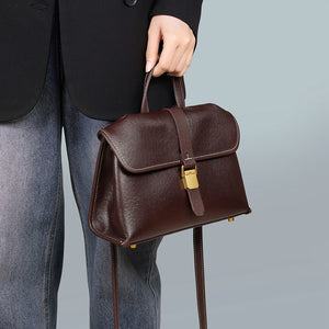 Woman holding the coffee brown flap satchel by its top handle, gold-tone buckle on the front, strap hanging down