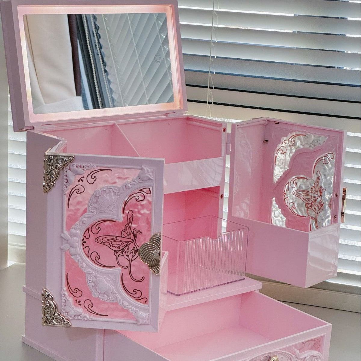 Open pink makeup cabinet with a lit mirror in the raised lid, a door open on a clear divider box, drawer pulled out