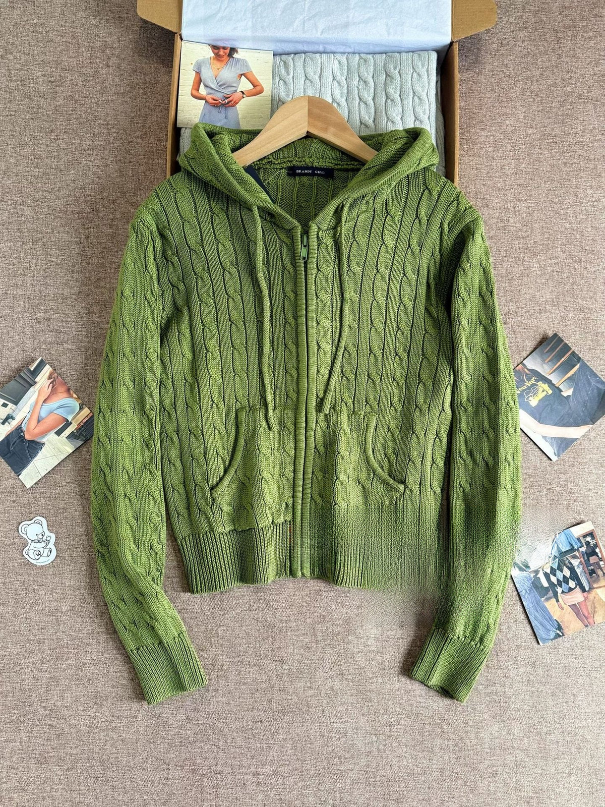 Avocado green cable knit hooded zip cardigan laid flat on a hanger with ribbed hem and front pocket