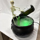 Skeleton holding a tilted potion bottle that pours a green stream into a black bat cauldron with rising mist