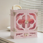 Pink makeup organizer cabinet with ornate double doors and a bow-shaped drawer pull on a white desk by a window