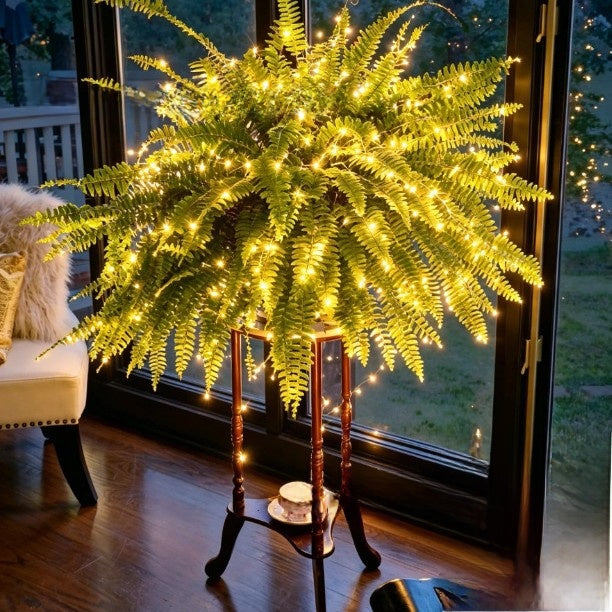 Lit artificial Boston fern on a tall wooden plant stand beside a window in a warm room