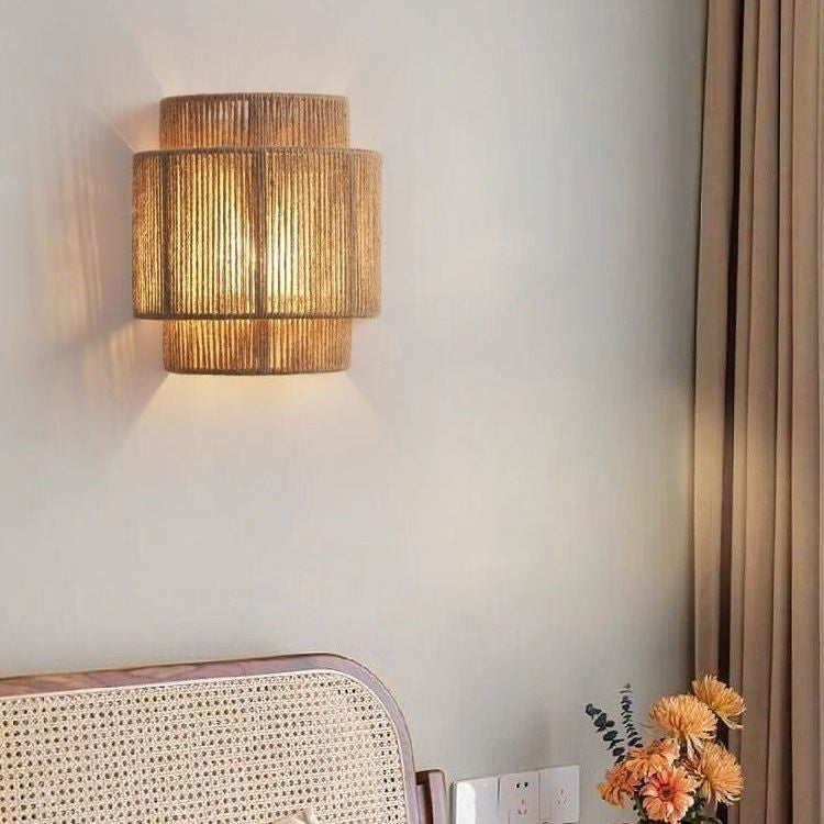 Lit hemp rope wall lamp with stacked tiers on a white wall above a cane chair back and orange flowers
