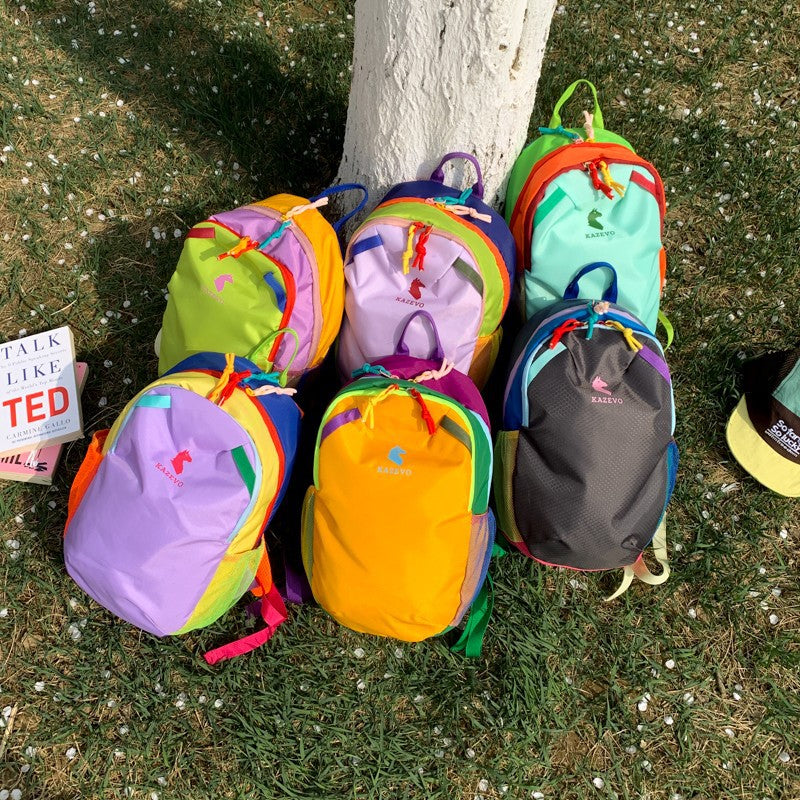 Six color block travel backpacks in purple, yellow, green, lilac, mint, and black lined up on grass