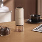 Cream electric coffee grinder with a top display disc beside a jar of coffee beans, a black cup and a newspaper