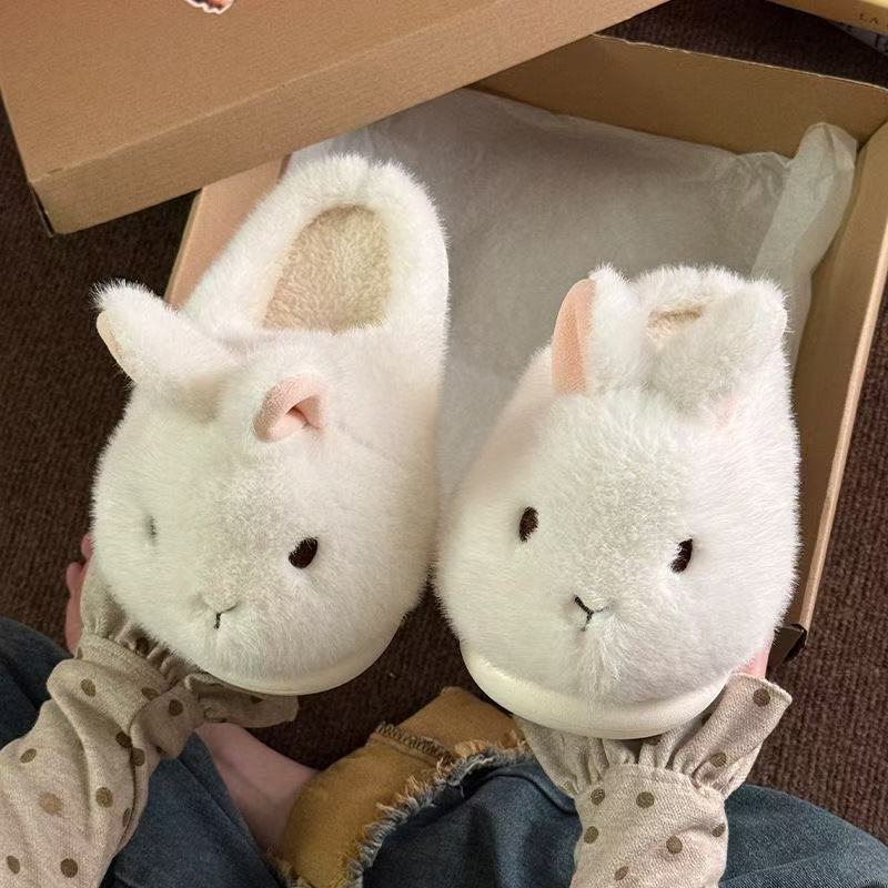 Two white bunny plush slippers with pink ears sitting inside an open cardboard box on a floor