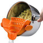 Orange silicone clip-on pot strainer on a stainless steel pot draining pasta, water pouring from the spout
