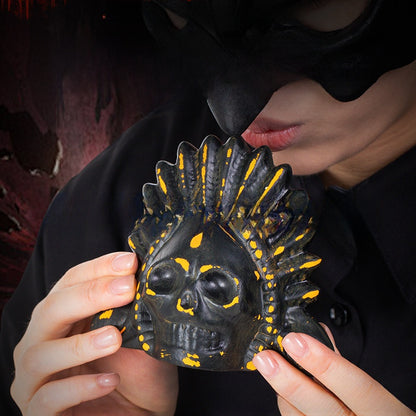 Person holding a black ceramic skull death whistle with orange feather accents to their lips on a dark background