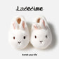 Pair of white bunny plush slippers with pink inner ears seen from above on a light panel with script lettering, view 3 of 3