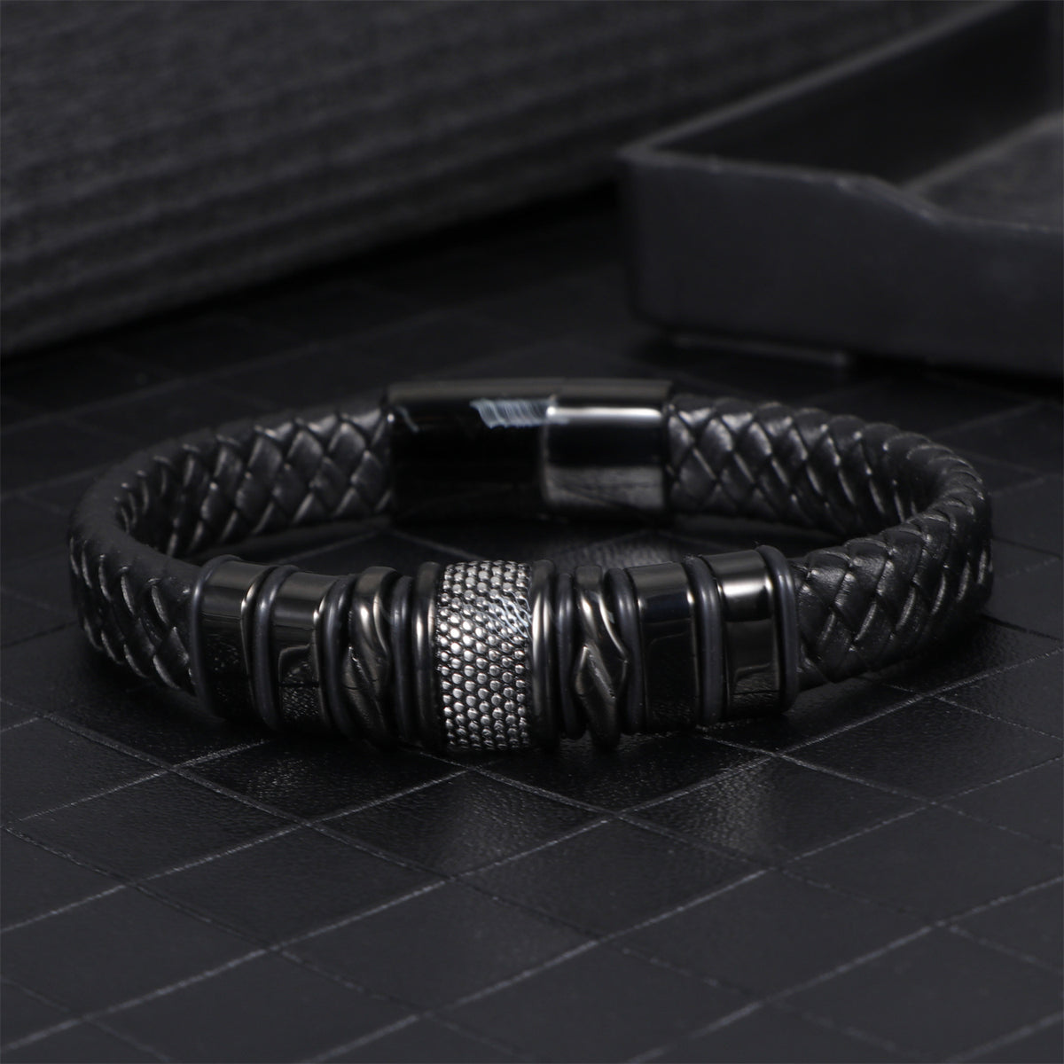 Mens black leather bracelet on a dark tiled surface beside a jewelry tray