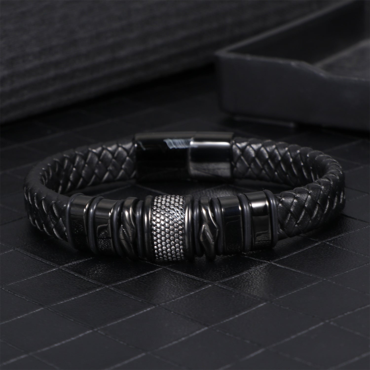 Mens black leather bracelet on a dark tiled surface beside a jewelry tray