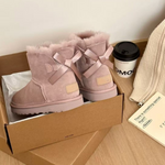 Pink sheepskin ankle boots with satin ribbon bows in a cardboard box beside a coffee cup and a knit scarf