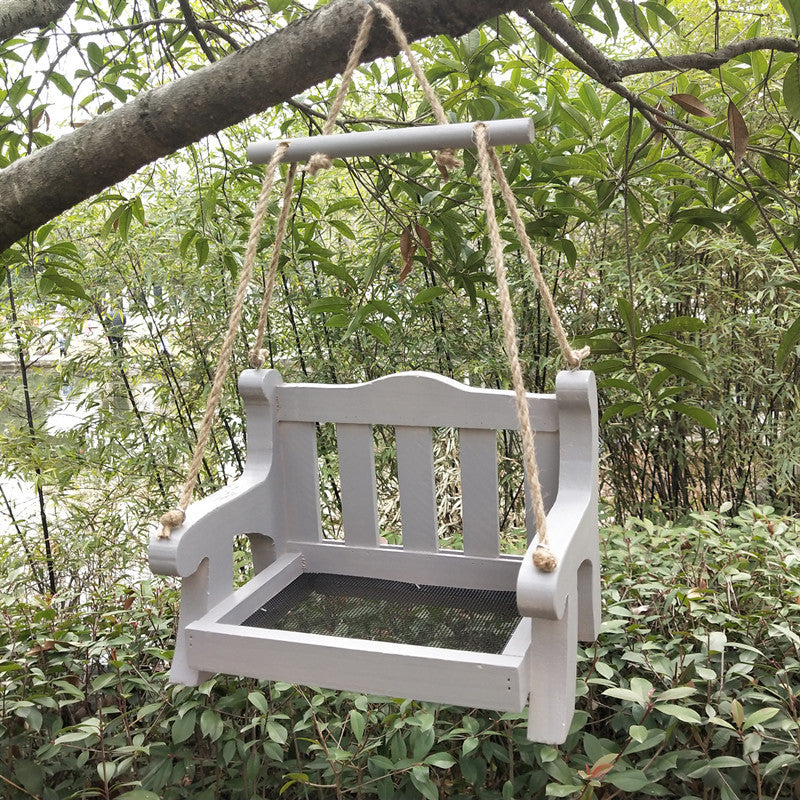 Light gray wooden bench bird feeder with a mesh floor and a gray crossbar hanging in a green garden