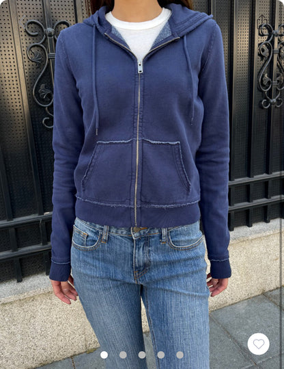 Woman wearing a navy zip-up hooded sweatshirt with raw edge seams and a front pocket, styled with jeans