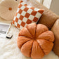 Orange eight-petal pumpkin pillow in front of a checkerboard pillow and a beige pleated round pillow on a sofa