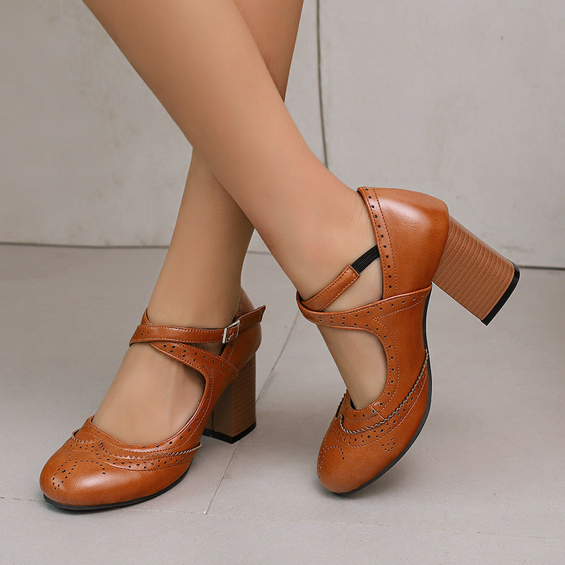 Caramel brogue Mary Jane block heel pumps with a buckled cross ankle strap, worn with legs crossed on a gray floor