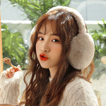 Woman wearing taupe beige plush winter earmuffs, side view, in front of green plants