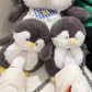 Dark gray plush penguin slippers worn on feet beside a large penguin plush toy with a gingham bib
