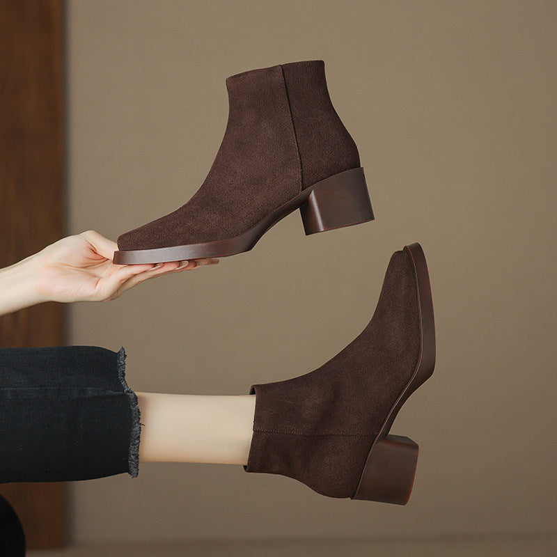 Brown suede finish ankle boots with chunky heel held in a hand beside a boot worn with jeans
