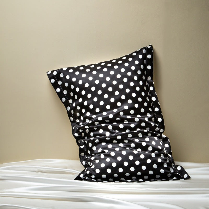 Black silk pillowcase printed with white polka dots, leaning on a beige wall above white silk