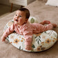 Baby lying on its tummy on a U-shaped nursing pillow with an orange floral cover on a woven rug