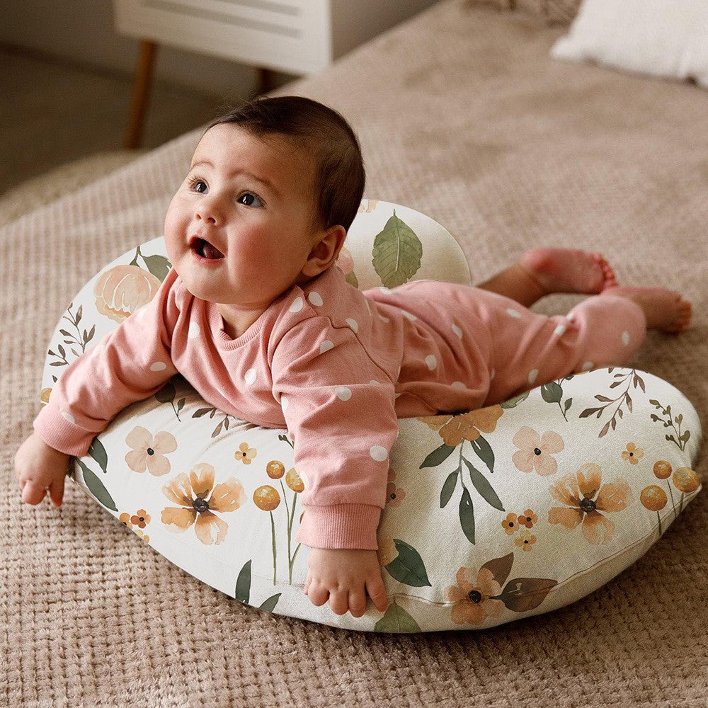Baby lying on its tummy on a U-shaped nursing pillow with an orange floral cover on a woven rug