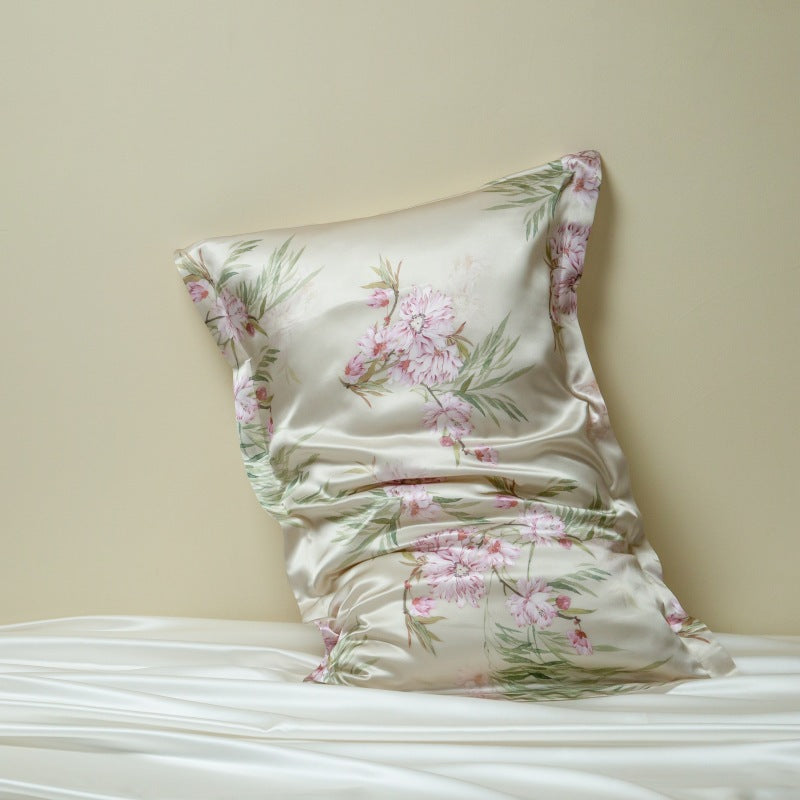 Cream silk pillowcase printed with pink blossoms and green stems, leaning on a cream wall above white silk