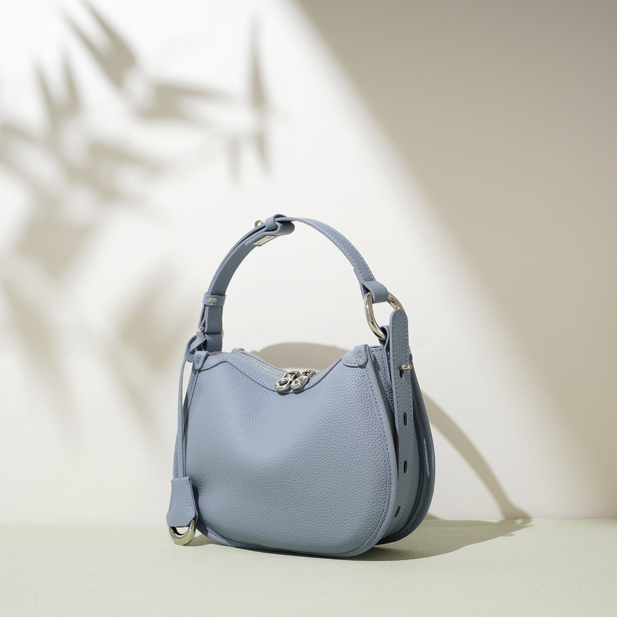 Dusty blue pebbled cowhide crescent bag at an angle with silver tone zip pulls, side ring and leather tab