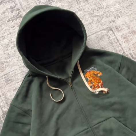 Close view of a green hoodie hood and zipper with an embroidered tiger on the chest crossed by a cream drawcord
