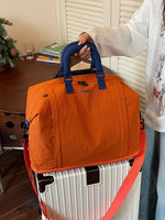 Orange Oxford travel duffel bag with padded handles resting on a wheeled suitcase