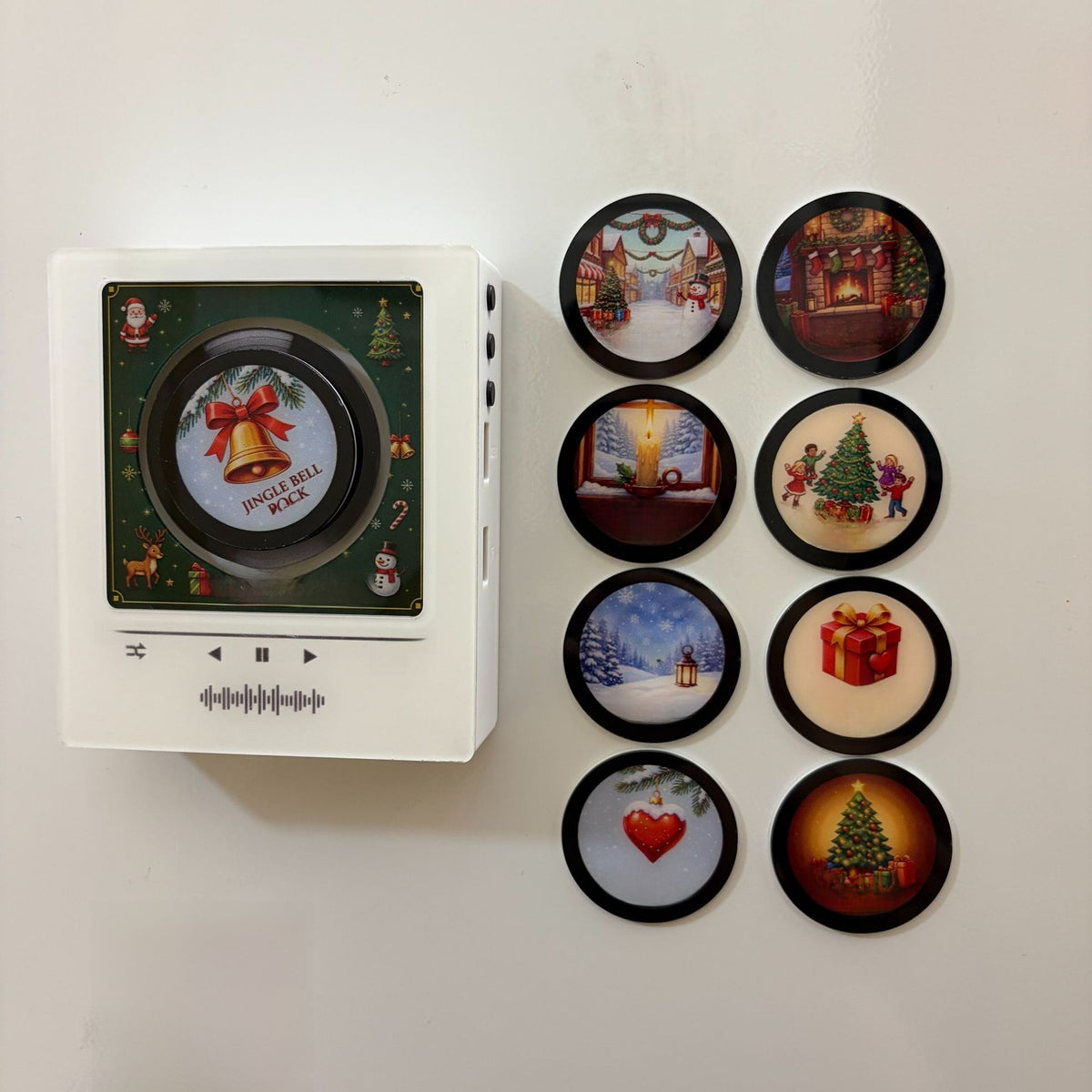 White NFC music player stuck to a wall with a green Christmas scene and eight round discs beside it