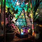 Bohemian diamond shaped projection lamp lit with colorful floral patterns beside houseplants
