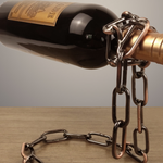 Dark wine bottle balanced on a copper toned iron chain loop rising from a coiled chain base on a wooden table