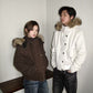 Man in an off white hooded jacket and woman in a dark brown hooded jacket with fur trim, standing beside a wooden bench