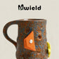 Brown and blue speckled ceramic mug with its handle on the left and an orange climbing hold decoration, side view