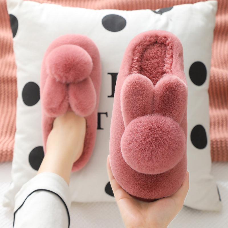 Burgundy plush bunny slippers with pom pom tails, one held in a hand and one worn on a foot over a polka dot cushion