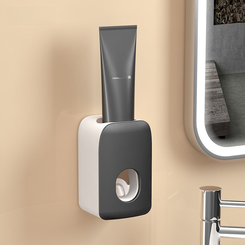 Gray wall mounted toothpaste squeezer holding an upright tube above the oval paste outlet in a bathroom