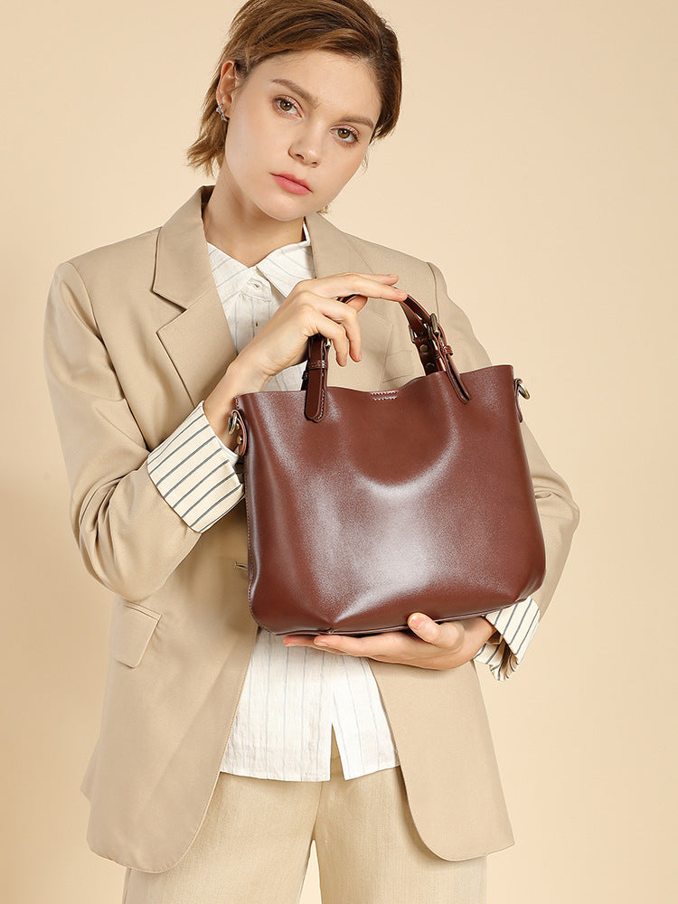 Woman holding a chestnut brown cowhide leather bucket tote by its top handles, wearing a beige blazer
