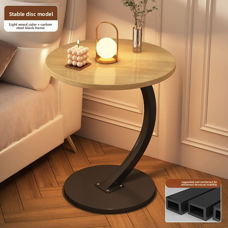 Light natural wood round top side table with a black steel C frame and disc base beside a bed, option Light Natural Wood